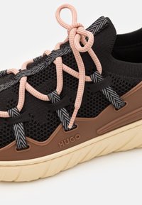 Schwarzer Sneaker aus Mesh mit brauner Gummisohle, pink-schwarz gemusterten Schnürsenkeln, ausgeprägter Nahtführung und Logo an der Seite.