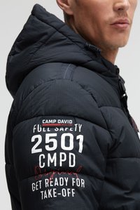 Man draagt een zwarte Camp David pufferjas met capuchon en witte tekst "2501 CMPD Full Safety Get Ready for Take-Off" op de mouw.