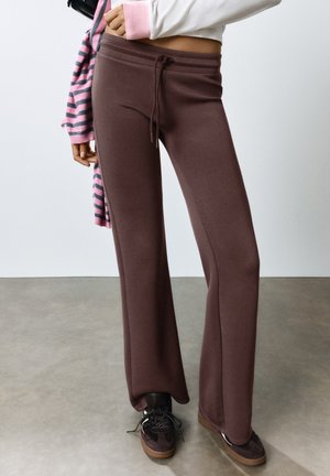Personne portant un pantalon large marron avec cordon, des baskets noires, un top court blanc et un pull rayé rose et gris drapé sur une épaule.
