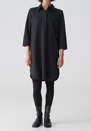 Černé šaty košilového střihu s dlouhými rukávy, límcem, zapínáním na knoflíky vpředu, dvěma náprsními kapsami, zaobleným lemem, nošené s černými legínami a kotníkovými botami.
