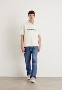 Bjela polo majica s logom "calvin klein", plave traperice, svijetlosmeđi tenisice; model stoji ispred neutralne pozadine.