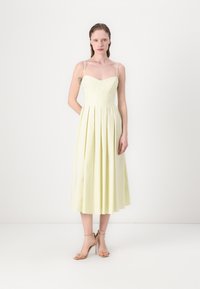O rochie midi galbenă cu decolteu în formă de inimă, bretele subțiri și fustă pliseată. Purtată cu sandale cu toc nud. Finisaj de material neted.