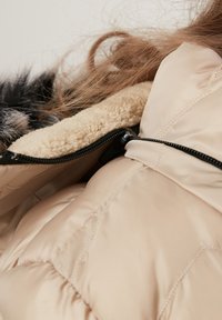 Beige termojakke med blød, lammeskindforing. Har en sort lynlås og kviltet design, der viser en glat, skinnende overflade.