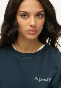 Sudadera azul marino con cuello y puños acanalados, que presenta un logo gris de "Superdry" en el pecho. Textura de tela suave.