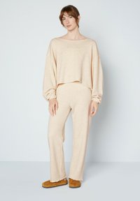 Beige fleece crop top en bijpassende broek met een ontspannen pasvorm. Zachte textuur met een licht rafeligezoom. Bruine slippers maken de outfit af.