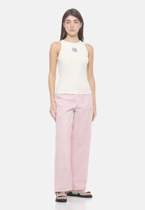 Donna in piedi che indossa un top bianco senza maniche con logo, pantaloni rosa larghi e sandali neri su uno sfondo bianco uniforme.