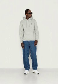 Man in grijze hooded sweatshirt, blauwe jeans, witte sneakers en zwarte zonnebril staat tegen een effen lichte achtergrond.