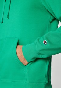 Felpa verde realizzata in morbido cotone, con tasca a marsupio e polsini a coste. Logo distintivo in rosso e nero sulla manica.