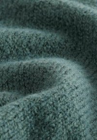 Texturiertes grünes Strickgewebe, das eine weiche, flauschige Oberfläche mit einem variierenden Stichmuster aufweist, das visuelle Tiefe und Wärme schafft.