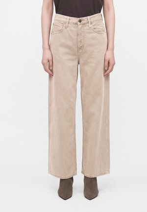 Personne portant un pantalon beige taille haute à jambes larges et des bottines foncées à bout pointu, se tenant devant un fond clair uni.