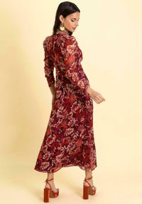 Rotes florales Midikleid mit langen Ärmeln, tailliertem Oberteil und fließendem Rock. Akzentuiert mit transparentem Stoff und kombiniert mit orangen Blockabsatzsandalen.