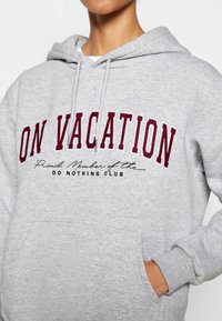 Personne portant un sweat à capuche gris clair avec un texte bordeaux « EN VACANCES » et un texte noir plus petit « Membre fier du CLUB DU RIEN FAIRE », main dans la poche.