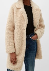 Manteau en fausse fourrure beige avec un col, extérieur texturé, design double boutonnage et poches latérales, porté sur un pull sombre et un jean.