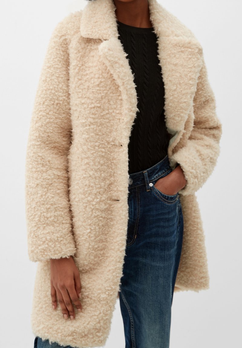 Manteau en fausse fourrure beige avec un col, extérieur texturé, design double boutonnage et poches latérales, porté sur un pull sombre et un jean.
