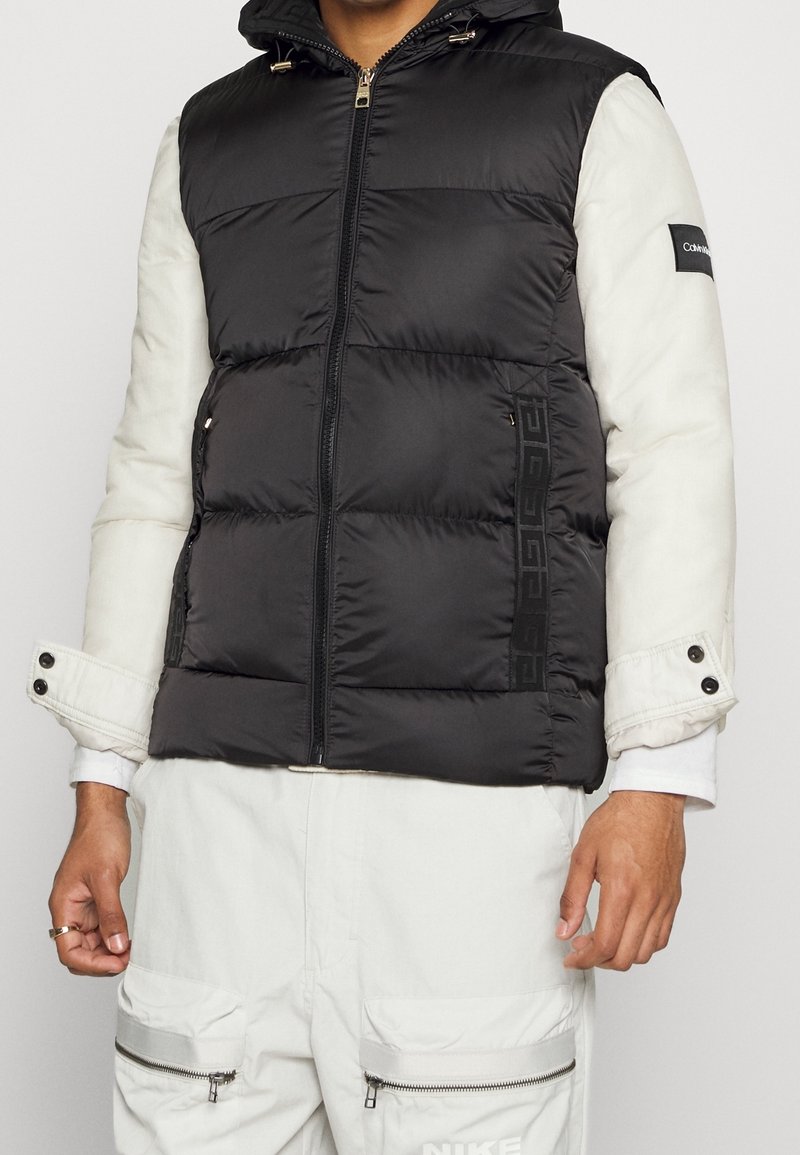 Gilet rembourré noir avec fermeture éclair à l'avant, capuche et manches blanches. Présente des accents de logo et des poches zippées. Porté avec un pantalon cargo blanc.