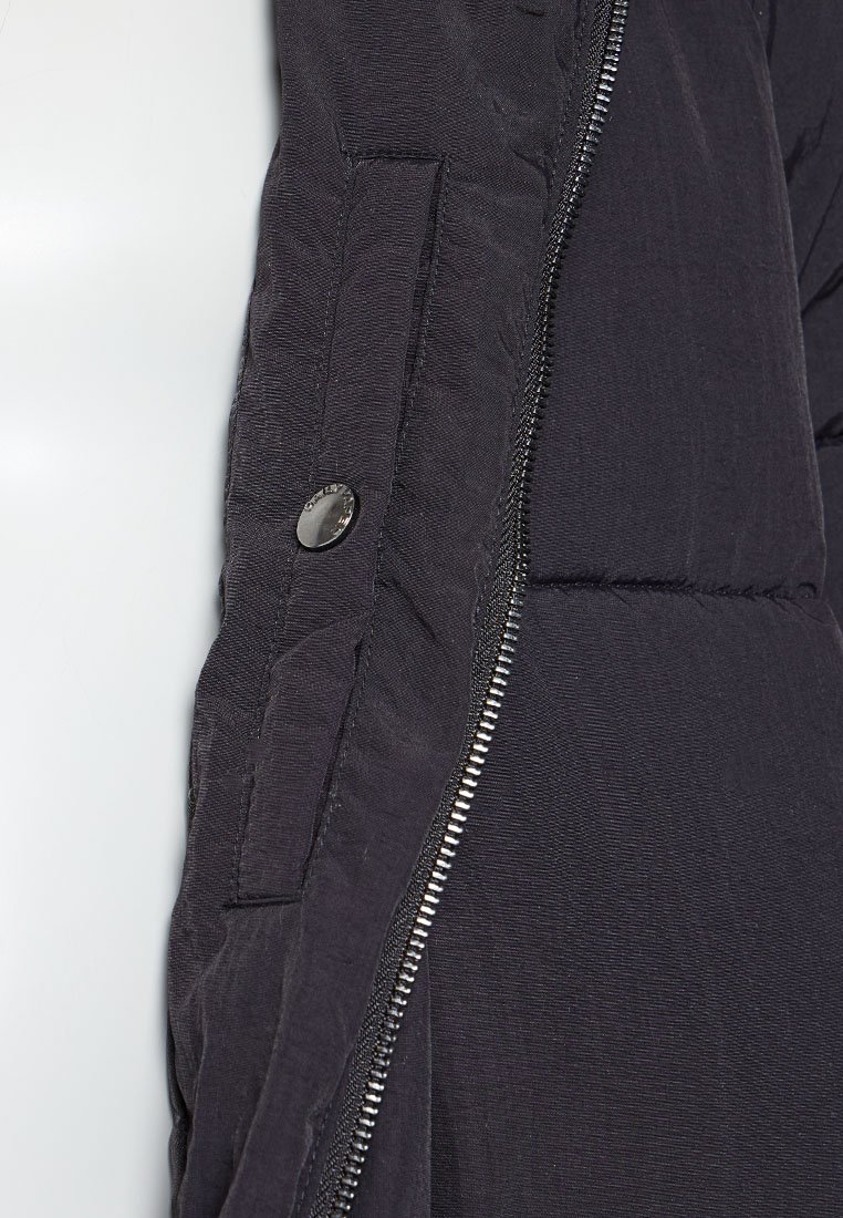 Sectie van een zwarte winterjas met een rits en drukknoop. Heeft een doorgestikte textuur en een gladde stof met een matte afwerking.