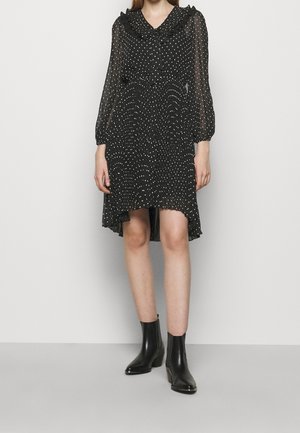 Femme portant une robe noire à manches longues avec des pois blancs et un col à volants, associée à des bottines noires, debout contre un mur uni.