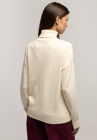 Sweter w kolorze kremowym z golfem, wykonany z dzianiny, z prążkowanymi krawędziami i luźnym krojem. Widoczne szwy na ramionach i plecach.