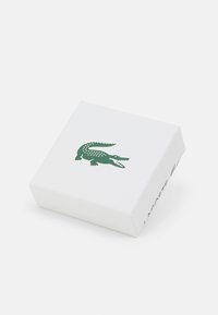 Balts taisnstūra kaste ar tekstūras virsmu, kuras augšpusē ir zaļais krokodila logo un sānos ir drukāts "LACOSTE".