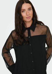 Crna košulja s punim korzetom, prozirnim dugim rukavima, ovratnikom i prednjim kopčanjem na gumbe. Meka, lagana tkanina.