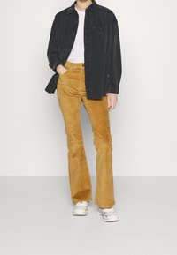Schwarze Cordhemd mit Knöpfen, darunter ein weißes Hemd, und senfgelbe Cordhosen mit Schlag. Graue Sneakers mit weißen Akzenten an den Füßen.