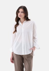 Camicia bianca con texture a pois, maniche lunghe e vestibilità rilassata, abbinata a pantaloni beige. Tessuto liscio, collo classico.