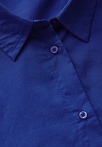 Camicia di cotone blu con colletto a punta, patta con bottoni e un bottone tondo viola, realizzata in tessuto liscio e testurizzato.