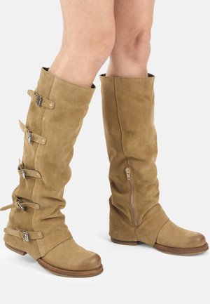 Knielange Stiefel aus beigem Wildleder mit niedrigen Absätzen, seitlichem Reißverschluss rechts und vier verstellbaren Schnallenriemen am linken Stiefel, getragen an nackten Beinen.