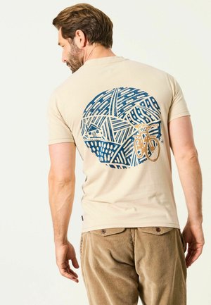Mann mit kurzen braunen Haaren, der ein beiges T-Shirt mit blau-orangefarbenem abstraktem Fahrraddesign auf dem Rücken und braune Cordhosen trägt.