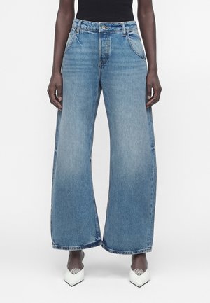 Personne à la peau foncée portant un jean bleu taille haute à jambes larges et des talons blancs pointus avec un détail en dentelle, debout devant un fond blanc.