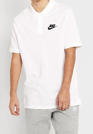 Weißes Poloshirt aus Baumwollmischung mit Zwei-Knopf-Leiste und schwarzem Nike-Logo auf der linken Brust. Kombiniert mit grauen Jogginghosen.
