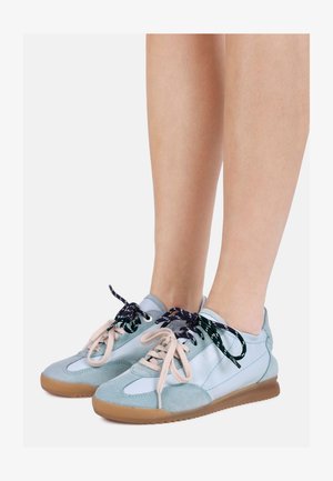 Piernas con zapatillas azul claro y gris con suelas de goma, cada zapato con cordones de colores negro y beige que no combinan.