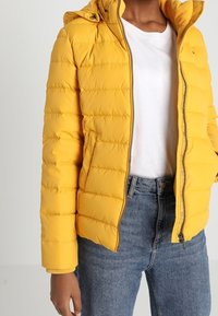 Żółta kurtka puchowa z zamkiem z przodu, kapturem i poziomym pikowaniem. W zestawie z białą koszulą i niebieskimi dżinsami.