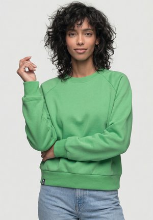 Frau mit lockigem Haar, die einen grünen Pullover und hellblaue Jeans trägt, steht mit einem Arm verschränkt und dem anderen erhoben.