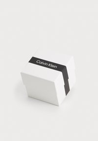 Boîte carrée blanche avec une bande noire arborant le texte "Calvin Klein". Texture lisse, design minimaliste, taille compacte.
