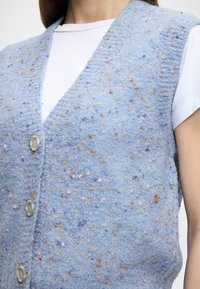 Lichtblauw vest gemaakt van gestructureerde stof, met kleurrijke vlekjes in roze, geel en oranje, voorzien van een V-hals en knopen aan de voorkant.