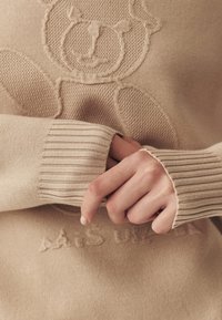 Maglione beige lavorato a maglia con design a rilievo di un orso, polsini a coste e un motivo testurizzato, con scritte del marchio visibili all'orlo.