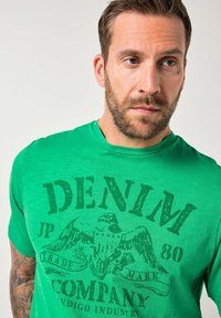 Groen T-shirt met korte mouwen gemaakt van zachte stof. Heeft een grote, donkergroene afbeelding van een adelaar en tekst met de woorden "DENIM JP 80 COMPANY."