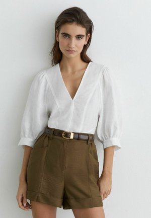 Młoda kobieta o długich brązowych włosach, ubrana w białą bluzkę z dekoltem w serek i bufkami na rękawach oraz oliwkowe, wysokowalne szorty z brązowym paskiem.