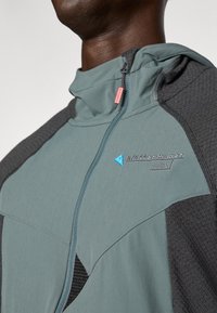 Mikina s kapucňou, ktorá kombinuje svetlomodré a tmavosivé materiály. Predný zips, textúrované akcenty, nášivka s logom a ružový ťahák na zips.