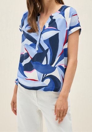 Kobieta ubrana w bluzkę z krótkim rękawem z abstrakcyjnymi wzorami liści w kolorach niebieskim, białym i czerwonym oraz białe spodnie, stojąca na neutralnym tle.