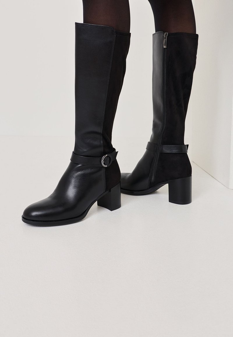 Botas de cuero negras hasta la rodilla con un zipper en el lado derecho, tacón grueso y una banda con una hebilla circular en el tobillo. Textura suave y mate.