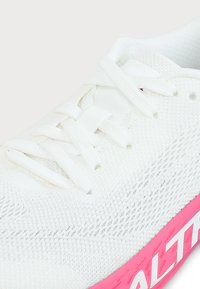 Chaussure de sport en mesh blanc avec des lacets blancs et une semelle rose comportant un texte blanc partiellement visible sur le côté.