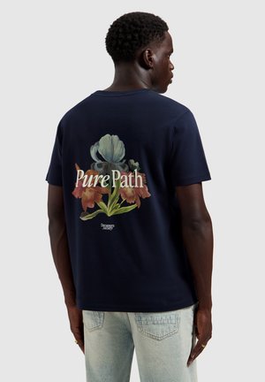 Man draagt een marineblauw T-shirt met een groot bloemmotief en de woorden "Pure Path" en "Dreamers Society" op de rug, gecombineerd met lichte spijkerbroek.