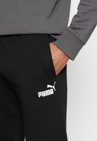 Šedá mikina Puma s elastickými manžetami, spárovaná s černými tepláky s bílým logem na stehně a bočními kapsami.