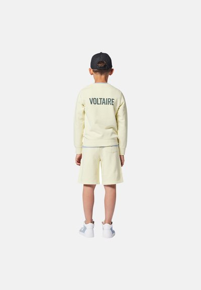 Sweatshirt jaune clair avec "VOLTAIRE" imprimé au dos, assorti de shorts. Il présente un col rond et des poignets côtelés. Baskets blanches et bleues.