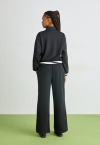 Giacca nera a maniche lunghe con colletto alto e polsini a righe bianche, abbinata a pantaloni neri larghi. Texture liscia, design casual.