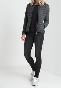 Puffet sort jakke med lynlås, lange ærmer og sidelommer, parret med en sort top og mørke jeans, accentueret af hvide sneakers.