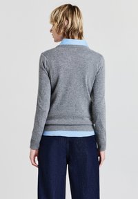 Pull en maille grise, avec un ourlet et des poignets côtelés. En dessous, une chemise à col bleu clair est visible. Pantalon large bleu foncé.
