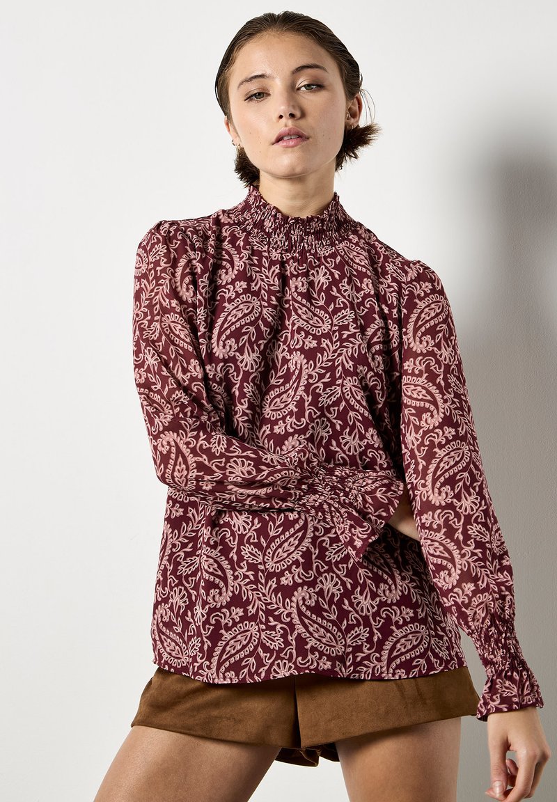 Blusa floreale bordeaux con maniche lunghe, scollatura arricciata e polsini smockati. Presenta un motivo paisley in rosa chiaro.